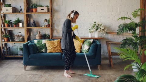 Attractive girl in headphones is listening to music and dancing wih mop during domestic work, she is mopping floor at home and having fun. Women, joy and houses concept.
