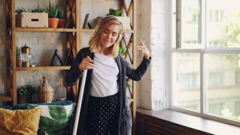 Attractive young lady is having fun during clean-up, she is singing and dancing with mop enjoying music in headphones. People, interiors and joy concept.