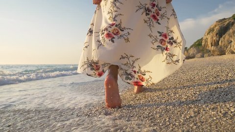 Slow motion, close up - Woman in a long dress walking barefoot in the water on the beach in the glow of golden light at sunset. Beautiful girl enjoying relaxing summer vacation