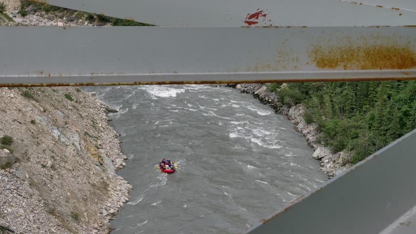 Red raft full of people going down a rapid in the river. Seen through a bridge metal structure in the foreground. Shot in 4K.