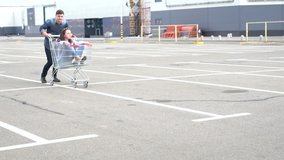 Happy smiling man pushing excited female friend in metal shopping cart across open parking lot beside building