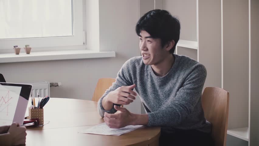 Young handsome happy Japanese man talking by the table, smiling at job interview in modern trendy light office. Royalty-Free Stock Footage #1015433821