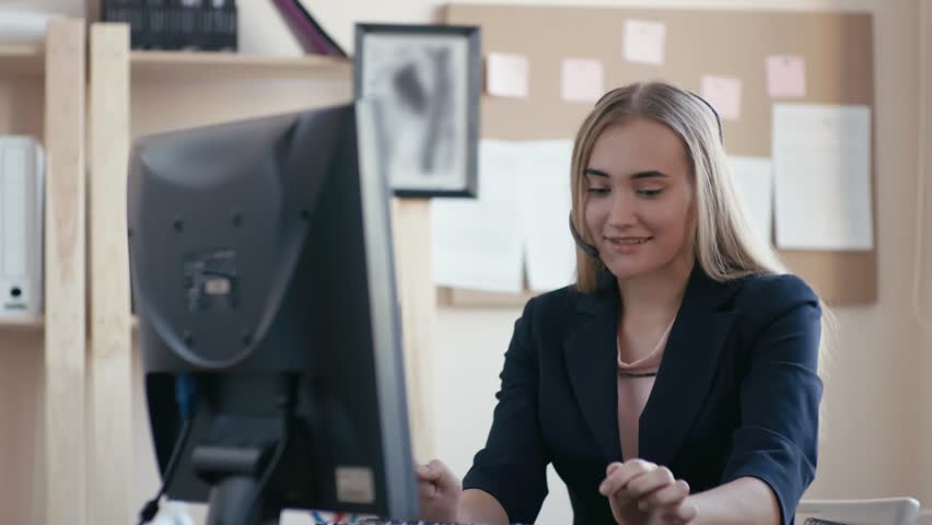 Call center employee at work in the office. A young girl sitting at the computer in the office. The girl takes a call on the phone, typing on the keyboard, looking at the computer screen and smiling. | Shutterstock HD Video #1015435168