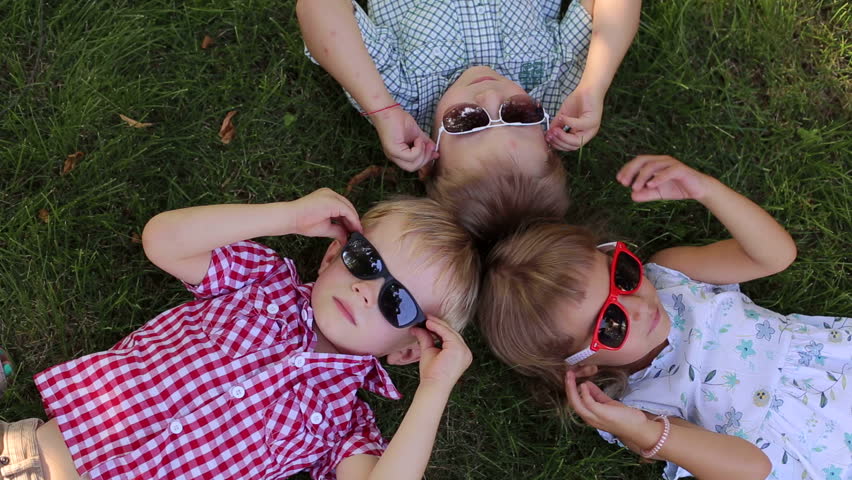 Three small children in sunglasses, two boys and one girl are lying on the grass head to head. Portrait of three small children in sunglasses in the Park. The view from the top.