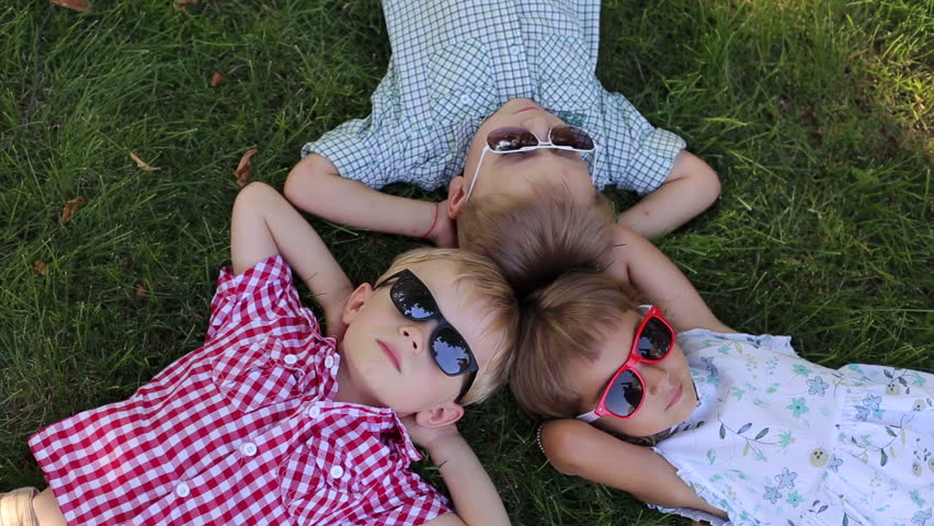 Three funny little children in sunglasses lay on the grass in the park, they laid their hands on the head. Portrait of three children in sunglasses in the park. View from above.