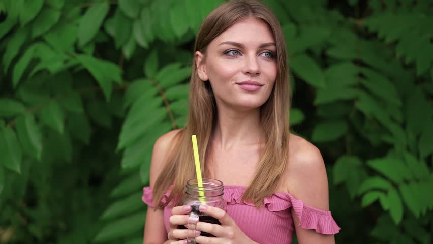 Attractive young woman in a pink dress is walking in a summer park and looking at camera. Seduction and flirting. Tracking slow motion medium shot