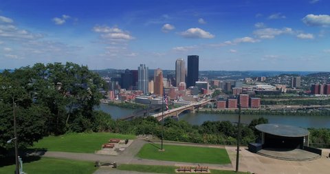 A wide aerial establishing shot of the iconic Pittsburgh, Pennsylvania skyline on a summer day as seen from Emerald View Park. American flag in the foreground.  	
