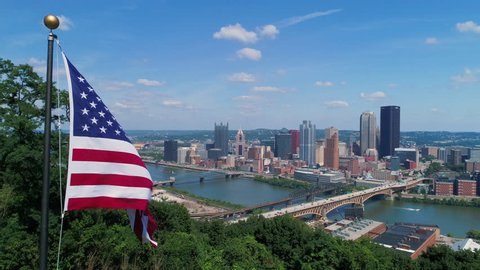 A slow push-in aerial view of an American flag on a flag pole above the Pittsburgh skyline on a summer day. As seen from Emerald View Park. Shot at 48fps for slow motion.  	
