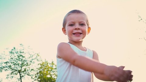 4-year-old boy in a white t-shirt laughs, covers his face with his hands, kid jumps up against the sky. Portrait of a cheerful active child on a nature background.