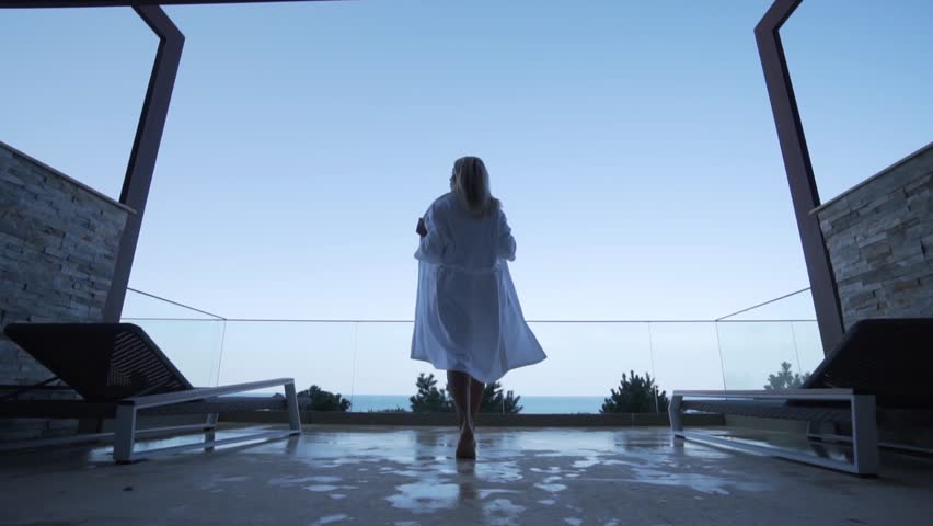 A blond girl in a white bathrobe walks out onto the balcony. Back view. Seaview. Royalty-Free Stock Footage #1015468048