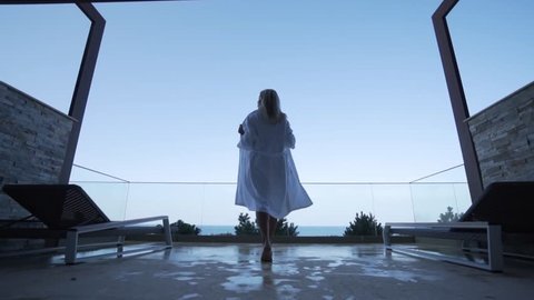 A blond girl in a white bathrobe walks out onto the balcony. Back view. Seaview.