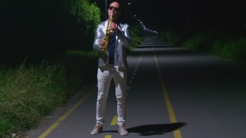 Fashionable saxophonist in a silver jacket and glasses plays at night on the road