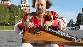 The minstrel in traditional Russian clothes plays an old Russian musical instrument gusli on the background of the Kremlin on red Square. Moscow, Russia
