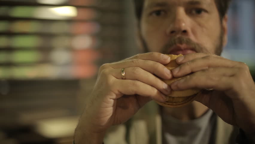 Caucasian bearded middle-aged man after work eating a hamburger in a summer cafe late at night on the background of the night city. | Shutterstock HD Video #1015484419