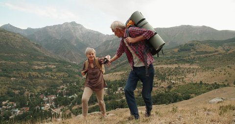 Mature caucasian senior on a hiking adventure taking wife's hand helping her climb up a mountain. tourism concept 4k
