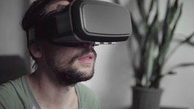 Young bearded hipster man using his VR headset display for watching the 360 video while sitting on sofa and eating cookies at home in the living room. VR Technology.