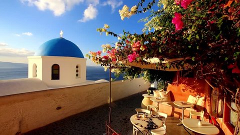 Blue church dome with wind playing with purple pink and white flower shrub on a terrace of traditional cafeteria in typical greek village of Oia, Santorini island, Greece.