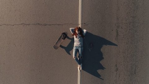 aerial. A teenager with a skateboard and lies on the roadway and listens to music on headphones. young girl hippy. View from above