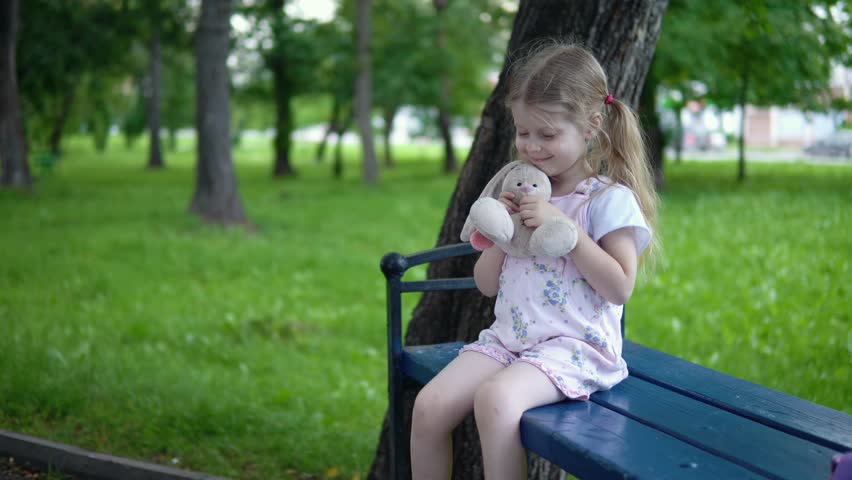 Little girl in pink overalls stroking a toy bunny, the child is showing a thumbs-up and sits on a bench in the park on a cloudy day.