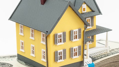 Toy model home sitting on top of a stack of 100 dollar bills USD, held in the palm of an adult male hand; HD ProRes clip