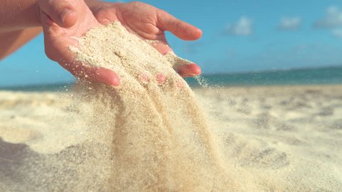 SLOW MOTION, CLOSE UP: Unknown young woman lets hot sand fall through her gentle fingers and back to the sunny coast. Cool slow motion of coarse particles of sand flying out female tourist's grasp.