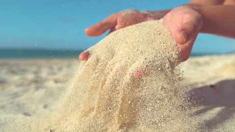 SLOW MOTION, CLOSE UP: Unknown female tourist having fun on empty exotic beach and playing with the soft sand. Carefree woman relaxing during her summer holiday by sifting sand between her fingers.