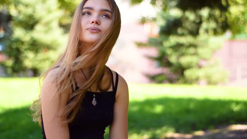 Attractive pretty young blonde woman with flying hair in the wind sitting in a bright sunset light. Woman in black dress sitting in parkland