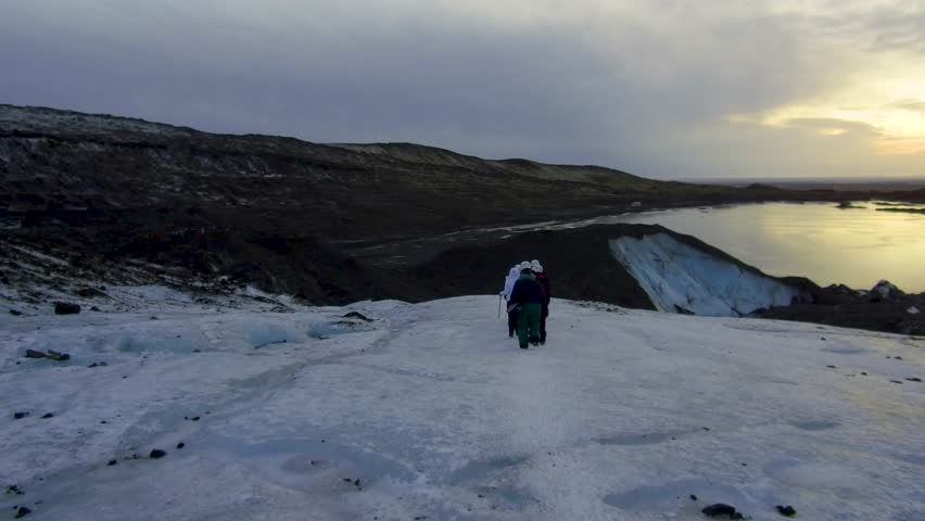 Dolly tracking shot camera follows a group of mountaineers hikers going down from a glacier on ice and snow in mountain toward foothills and a frozen lake during twilight