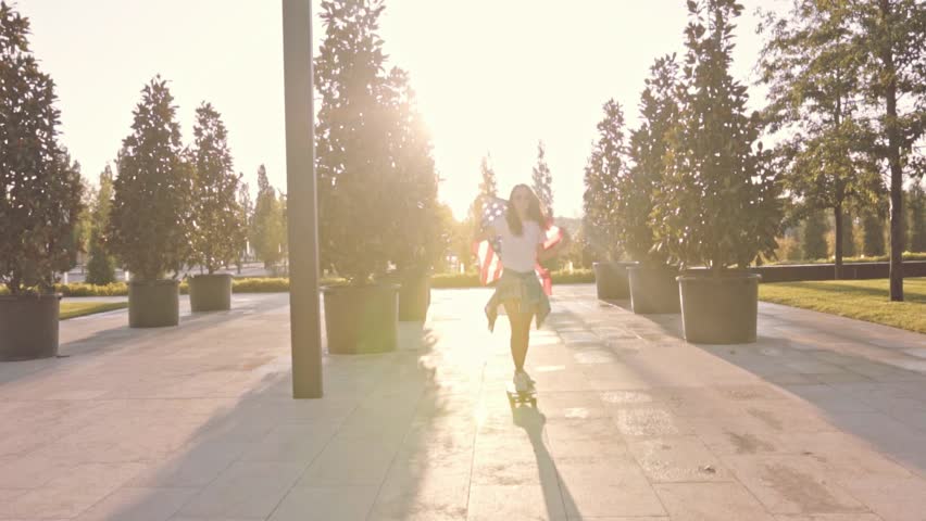 Free and careless young woman in pink t-shirt and denim shorts ride her longboard in urban park zone of the megapolis, proudly holds american flag of her country high in the air