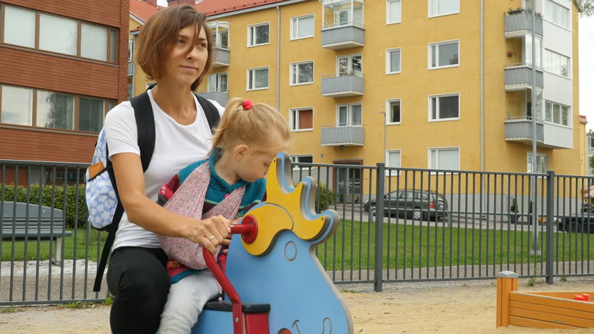 Happy family, mother with little child swinging on swing set at children playground. Mom playing with her daughter