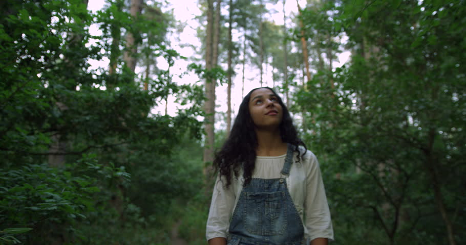 A Pretty Mix Race Young Student Girl Exploring The Woods on A Summers Day. Wearing Denim Dungarees and White Summer Shirt. Walking Through the Forest Looking Up at the Tall Beautiful Trees Smiling.