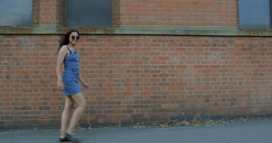 A Pretty Mix Race Young Girl Exploring The British Streets On a Summers Day.Wearing a Blue Denim Summer Dress With Trendy Sunglasses. Walking In front Of a Brick Wall. Having Fun On A Summers Day. | Shutterstock HD Video #1015553590