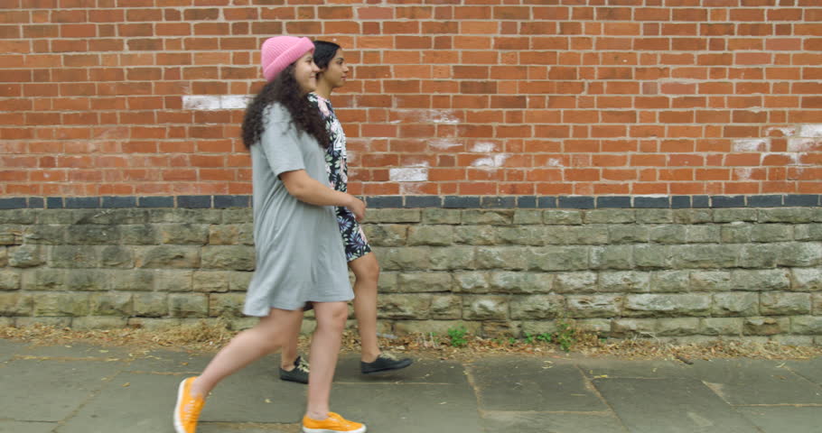 Two Pretty Mixed Race Young Girl Twins Walking Along a Street in Front of a Brick wall Cheerfully on a Summers Day. Wearing Pretty Summer dresses. Having Fun Together.