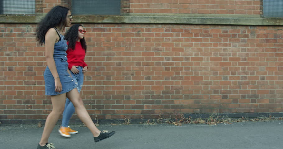 Two Pretty Mixed Race Young Sisters Walking Together Along a Street in Front of a Brick wall Cheerfully on a Summers Day. Wearing Blue Denim Clothes With Trendy Sunglasses. Exploring the City. | Shutterstock HD Video #1015557148