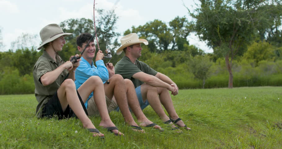 A father and sons sitting on a grassy riverside knoll having fun fishing.
