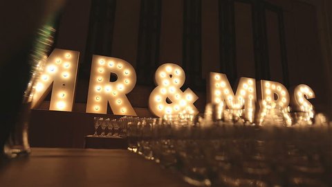 Retro lIghtbox theater style of text Mr & Mrs decoration on the backdrop in the wedding reception dinner night party with a group of wine glasses blurry in foreground, Low angle, Dolly slider scene.
