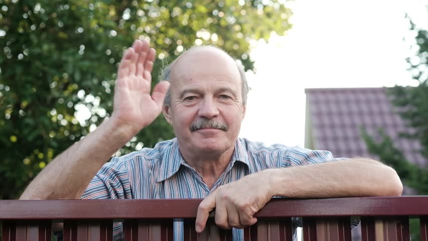 Friendly caucasian old man waving hi or farewell, isolated outdoors background with green trees and fence. Homeowmer saying goodbye to his guest Royalty-Free Stock Footage #1015589812