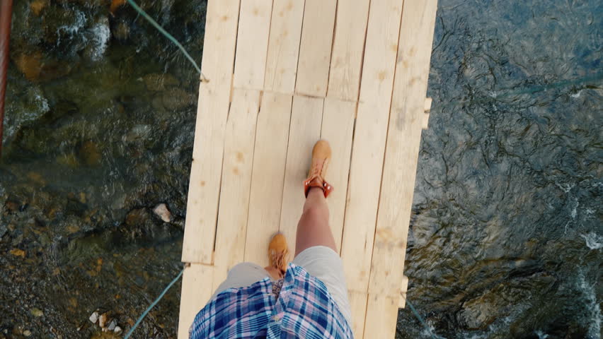 A woman walks along a narrow, shaky bridge across a mountain river. 