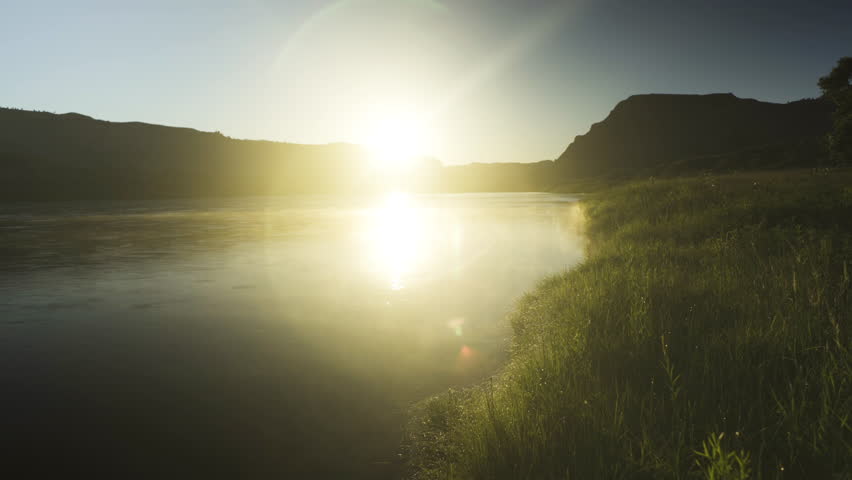Sun rising over the flowing river with dewey grass on banks in the badlands of Montana