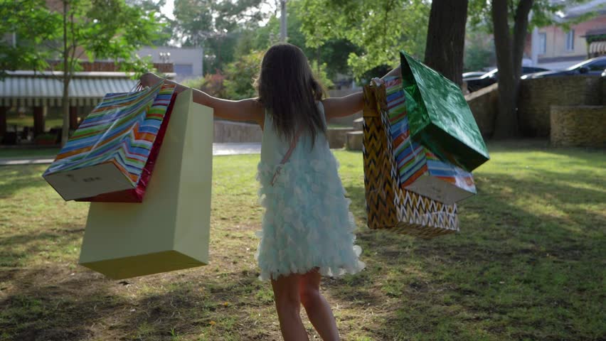 birthday, happy child girl with lots of gift bags in hands running to green grass in park