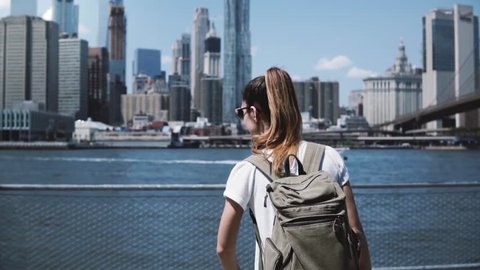 Back view of young happy tourist girl with backpack and arms wide open with joy at famous Manhattan skyline in New York