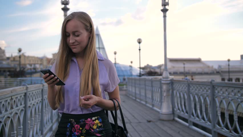 Portrait of young attractive blonde businesswoman walking on city bridge with smartphone in hand. Female xecutive texting and scrolling newsfeed on social media during a stroll at lunch break