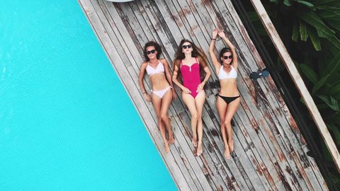 AERIAL. Camera zooming out. Top view of three young woman in sun glasses, lying near pool at luxury jungle villa. Three friends relaxing at the swimming pool.