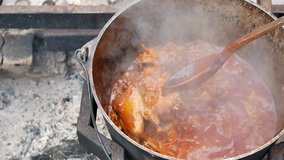 making soup pilaf in nature open fire while tourist hiking. stirring fried chiken with big wooden spoon at huge big cauldron. high angle shot