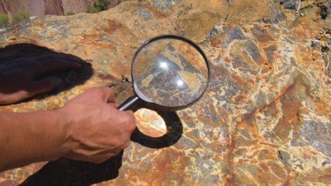 Geologist hand examines rock crystals mineral under magnifying glass. A large block of rock with inclusions of minerals lies in the mine dump. Study of mineral composition of minerals in ore: geology