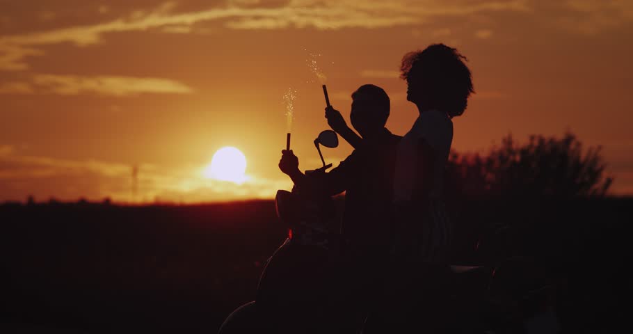 Amazing sunset beautiful couple stand behind the retro motorcycle happy holding sparklers looking to amazing nature view. slow motions. silhouette
