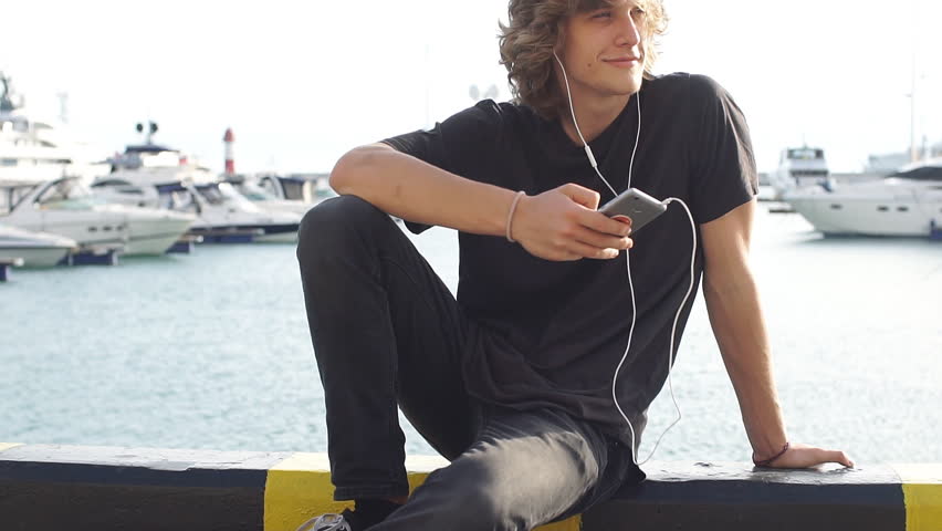 Young man using a cellphone while sitting at longboard on pier witth yacht. Slow motion