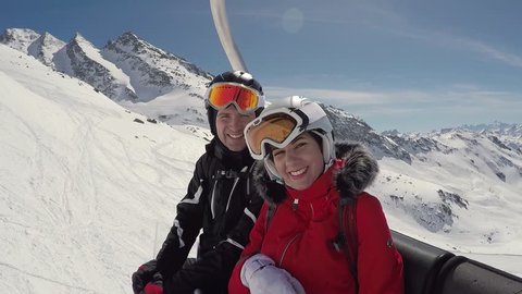 A loving couple of skiers climbs up the mountain on a ski lift, they have fun and are fooling, portraying funny faces on their faces, smile a lot and look at the camera