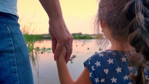 Father is standing with his small daughter near lake and watch ducks, holds her hand, sunset, family concept