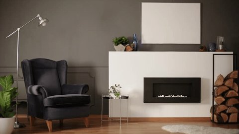 Metal lamp placed next to black armchair standing in dark sitting room interior with empty poster on wall, decor on shelf and eco fireplace in the video